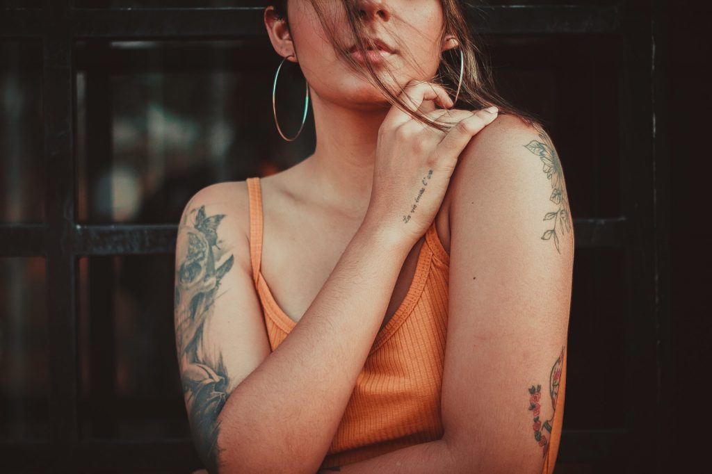 Das Bild zeigt eine Frau in orangenem Top mit Tattoos und großen Ohrringen.